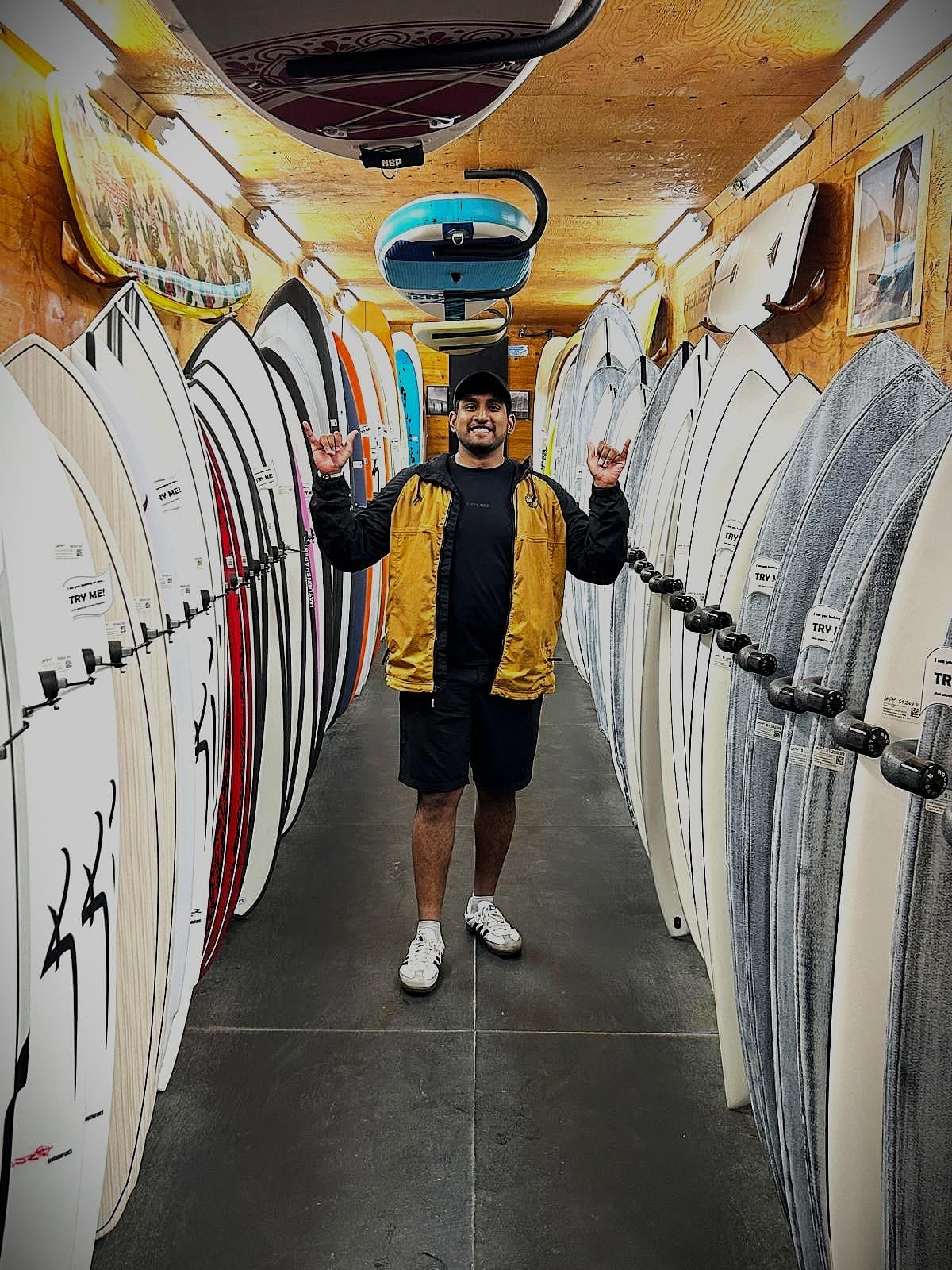 Rahil grinning between two full racks of surfboards in a shop