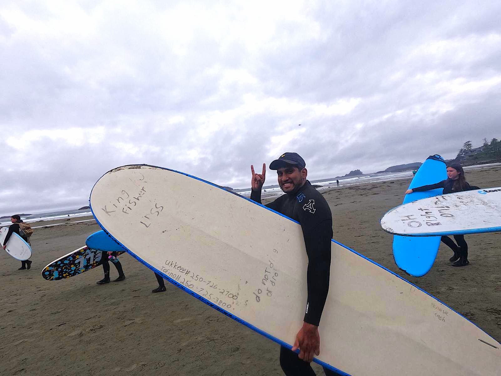 Rahil on the beach in Tofino, longboard under one arm, horns up